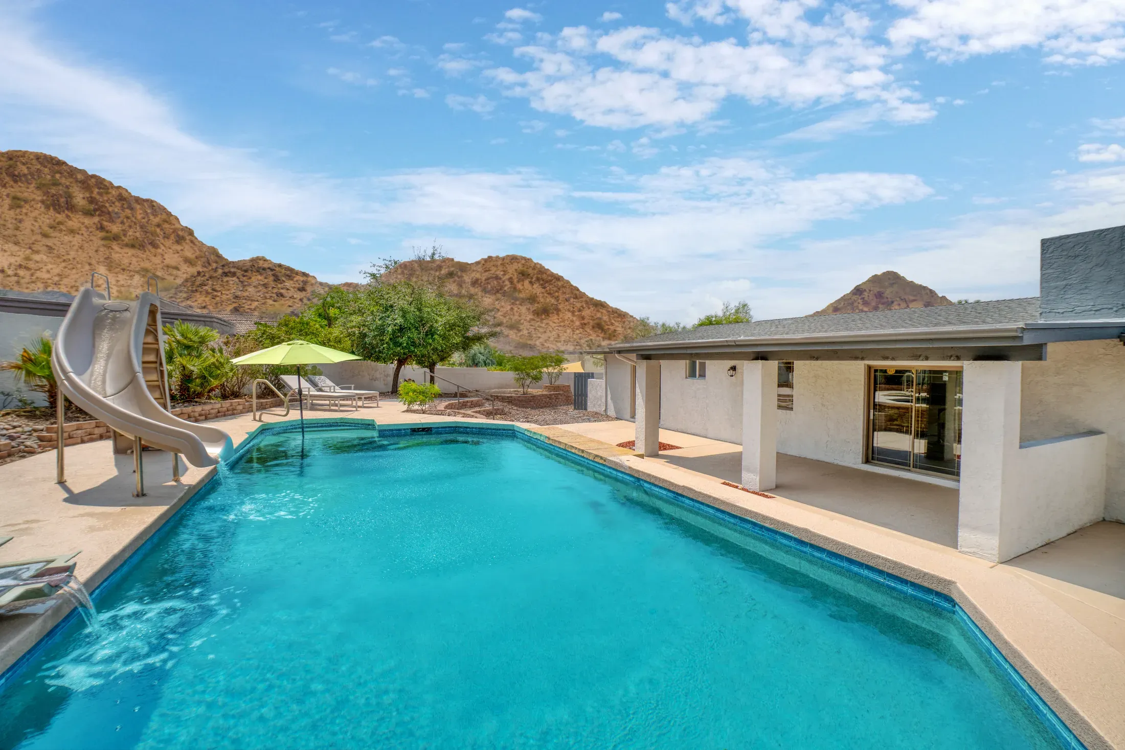 A backyard swimming pool with a slide at a desert home, mountains rising behind the patio